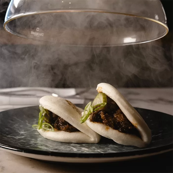 Two bao buns on a plate with glass lid, beside a steaming pot of meat at Beefbar Monaco, showcasing a delightful culinary presentation.