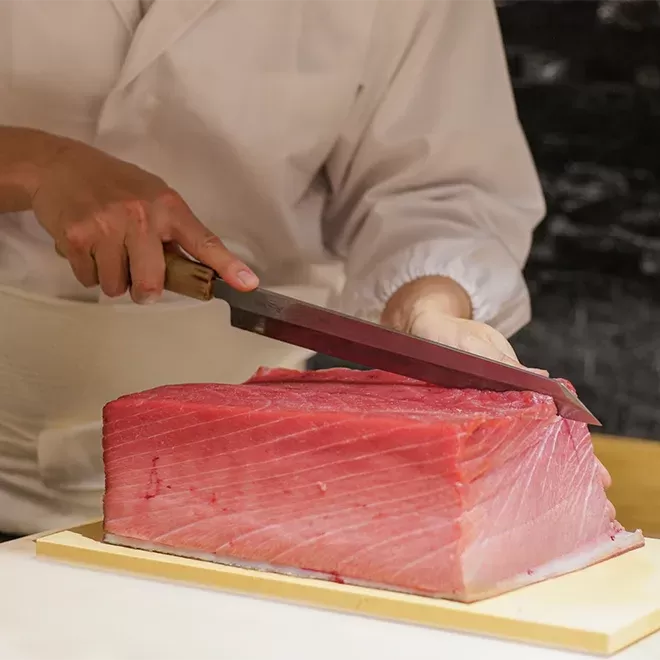 A chef in uniform skillfully cuts raw tuna on a cutting board at Taku restaurant, using a professional chef's knife.