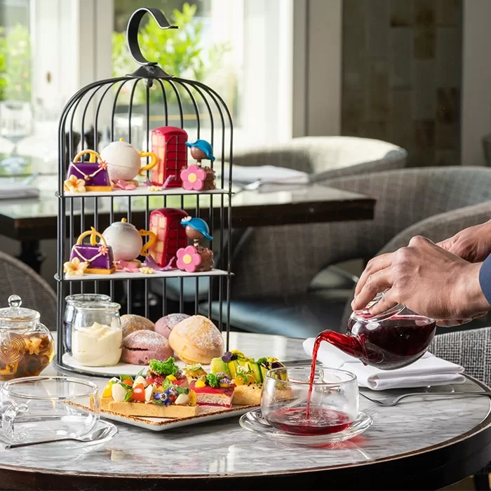 A cake stand with a mix of multi-coloured cakes and jars of clotted cream stands on a marble table with a man pouring a tea pot of red liquid into a cup.