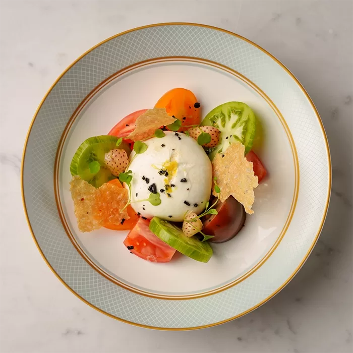 A plated dish featuring a round piece of burrata topped with black sesame seeds, surrounded by colorful heirloom tomato slices, crispy cheese shards, and microgreens on a decorative plate at Café Biltmore, Mayfair.