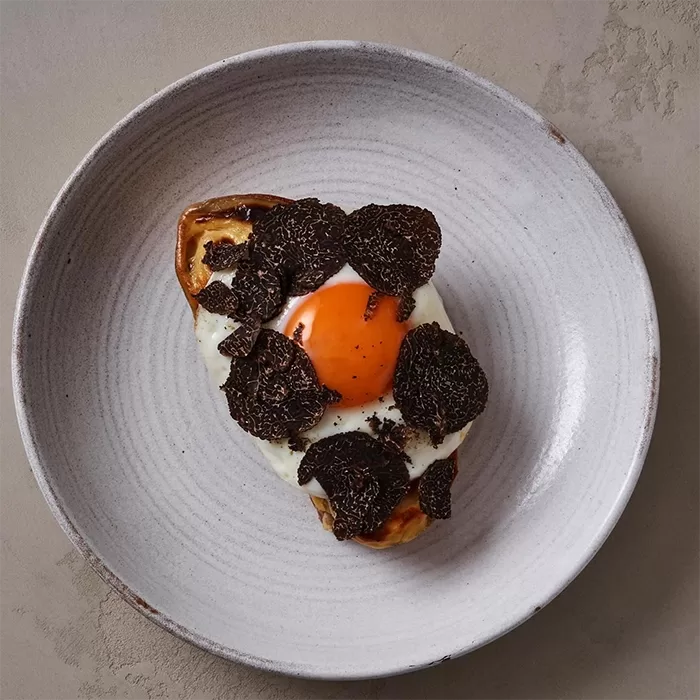 A gourmet dish featuring a fried egg on toasted bread with creamy sauce, topped with slices of black truffle, served on a white plate against a neutral background served at Hideaway, Mayfair.
