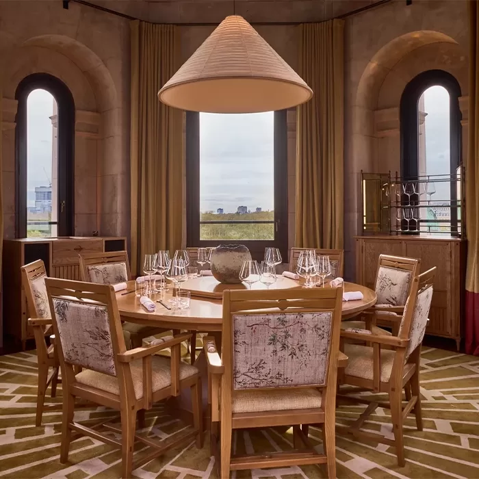 Elegant dining room with a round table set for six, featuring wooden chairs and a large overhead lamp. Two tall arched windows with curtains frame the background, offering a view of the cityscape. Decorative patterns adorn the carpet and chairs at Kioku by Endo: Turret, London.