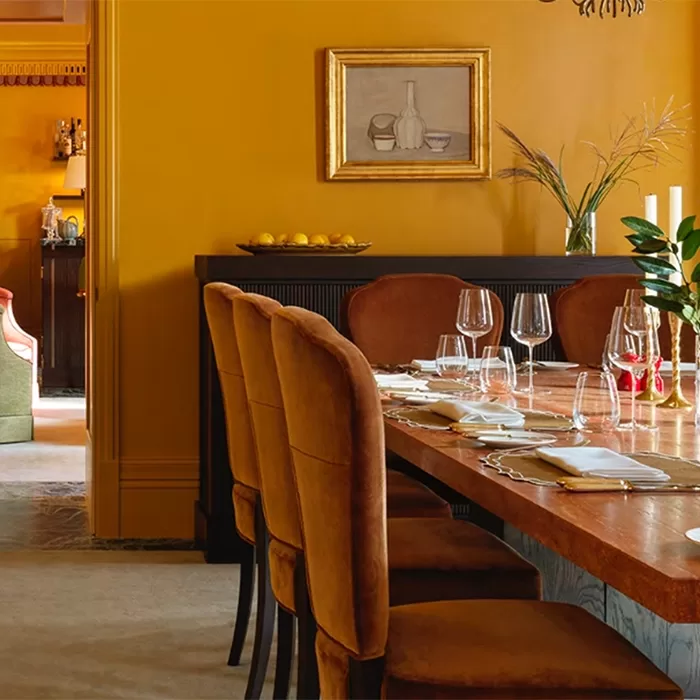 A dining room with mustard yellow walls features a long wooden table set with wine glasses and white napkins. Brown upholstered chairs line the table. A painting and a bowl of lemons are on a black console table in the background at Mount Street Restaurant: The Italian Rooms, London.