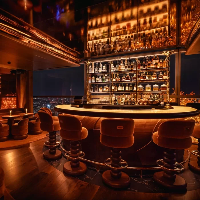 A cozy, dimly-lit bar with plush, brown, circular seating surrounding a glowing counter. Shelves stocked with various bottles are illuminated in the background, creating a warm, inviting ambiance. City lights are visible through large windows at SAMBA ROOM, London.