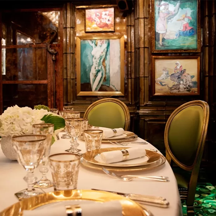 An elegant dining table set with gold-rimmed plates, glasses, and white napkins, surrounded by green upholstered chairs. The room features ornate wood paneling and colorful paintings on the walls, creating a luxurious atmosphere at Scott’s: Renoir Room, London.