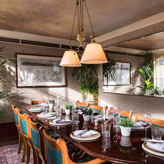 A warmly lit dining room with a long wooden table set for eight. Green upholstered chairs surround the table, which is adorned with plants and dinnerware. Large windows and wall art add to the cosy atmosphere at Chiltern Firehouse, London.