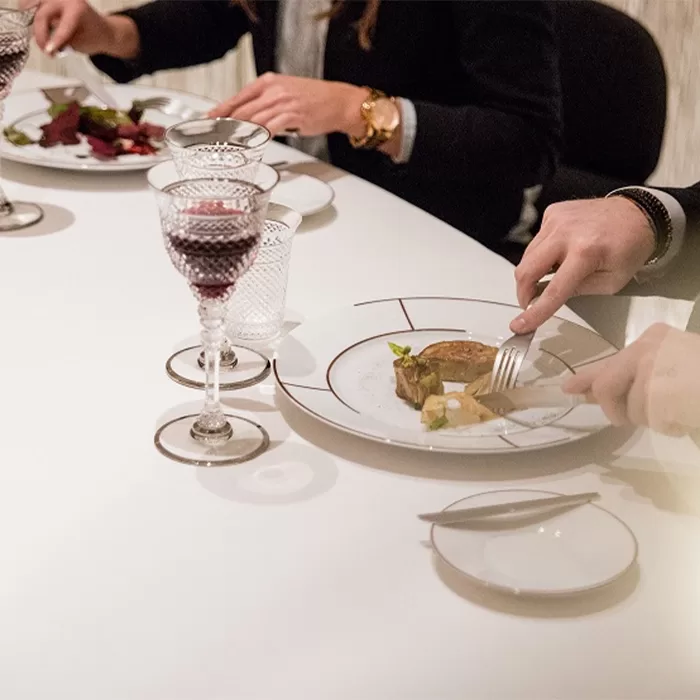 People dining at a white table set with wine glasses and elegant plates of food. A person cuts meat with a fork and knife, and another has a salad in front of them. The atmosphere appears formal and sophisticated at Alain Ducasse at The Dorchester: La Table Lumière.