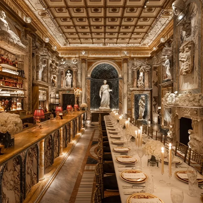 Luxurious dining room with marble walls and columns, adorned with classical statues and intricate ceiling designs. A long dining table with elegant place settings and tall candles stretches alongside a marble-top bar with red lamps and flower arrangements at Bacchanalia: Apollo’s Muse.