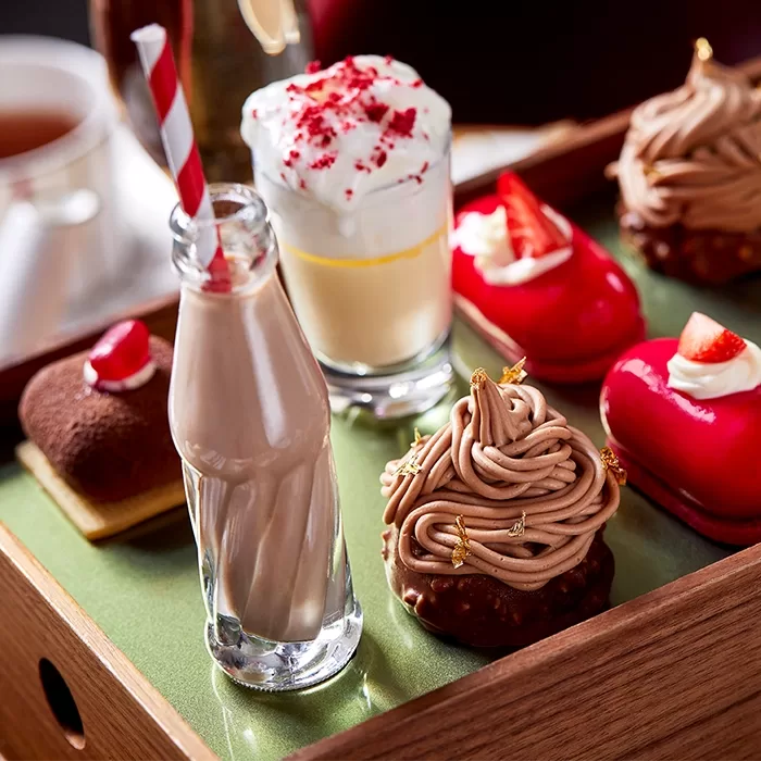 A wooden tray with a green bottom with a chocolate milkshake in a cola bottle with a red and white striped straw coming out. There is a vanilla milkshake next to this with whipped cream and strawberry sprinkles surrounded by small cakes.