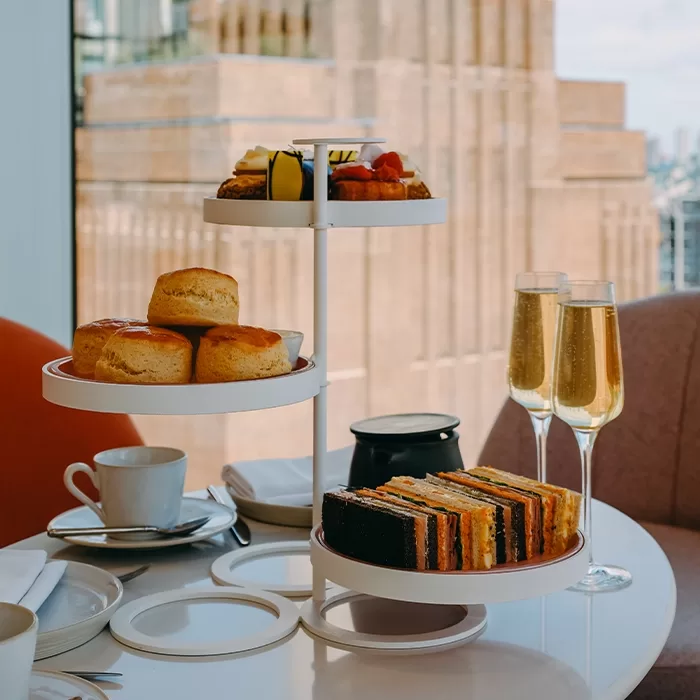 A table site next to a window overlooking a building. The table has 2 glasses of prosecco sat on it, next to a teapot and cup of tea. There is a cake stand with scones, cakes and sandwiches on 3 different tiers.