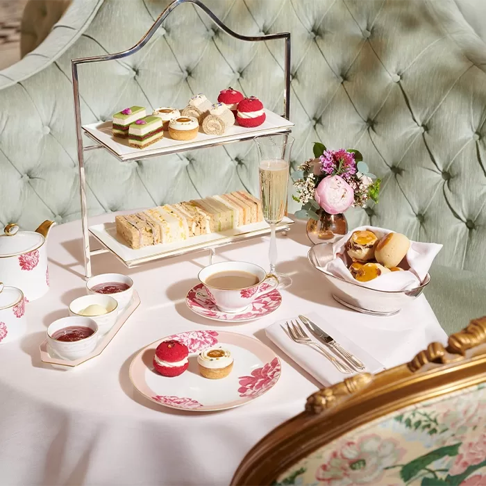 A white table cloth with white and pink plates laid on it full of cakes. A cake tray sits at the back of the table with 2 tiers, the bottom of lined finger sandwiches and the top a mix of different bite size cakes. On the table is jam, butter and a bowl of bread.