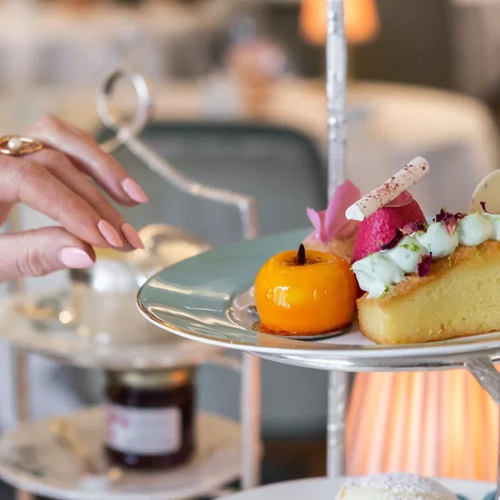 A close up on a lady's hands with pink nails reaching for a cake on a zoomed in cake tray showing some orange fruit and a slice of cake.
