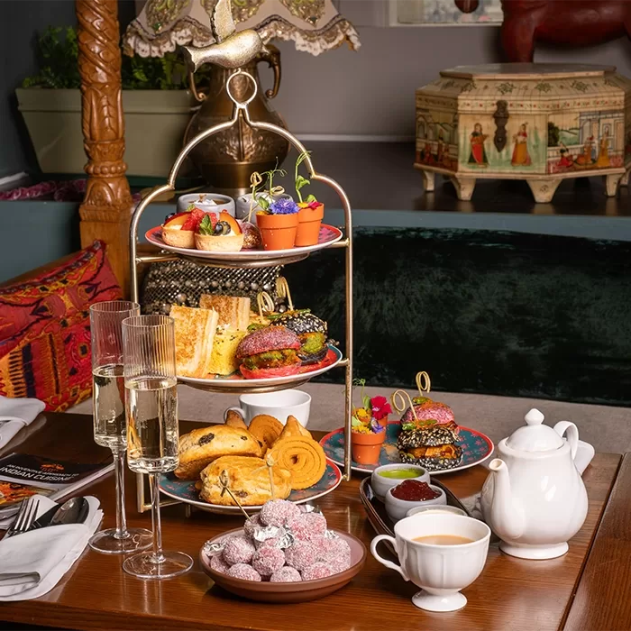 A wooden table with 2 glasses of prosecco, a China cup or tea and a teacup. There is also a 3 tier cake stand with pastries, clotted cream and a mic of coloured cakes, jam and butter,