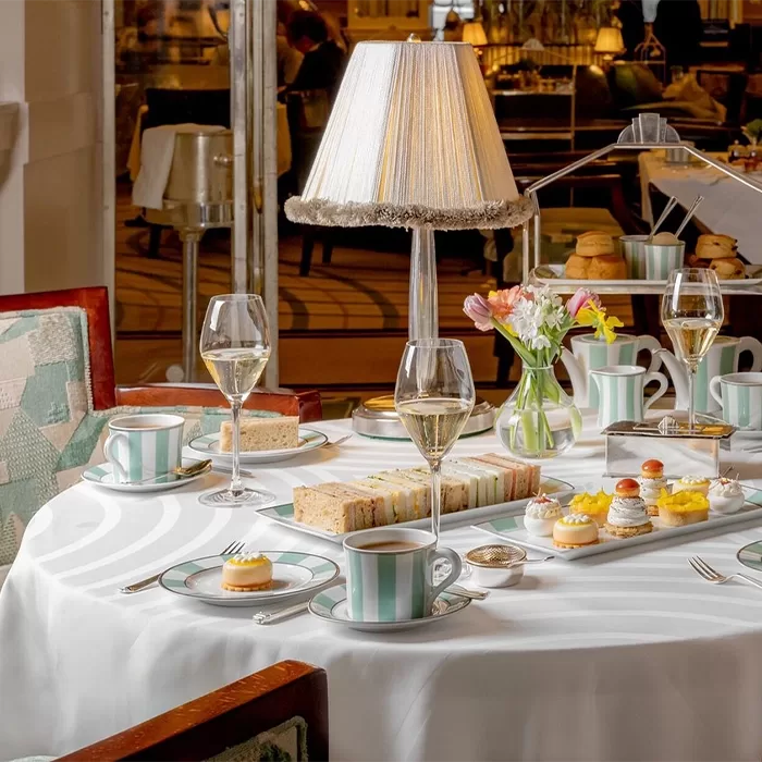 A table with a a tray of finger size sandwiches on it next to a plate of cakes and cups of tea and glasses of prosecco.