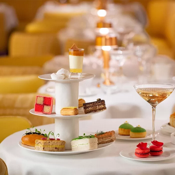 A white table with a white cake stand on it with a mix of finger size sandwiches and cakes. On the table is small white plates with red macaroons and a glass of rose wine.