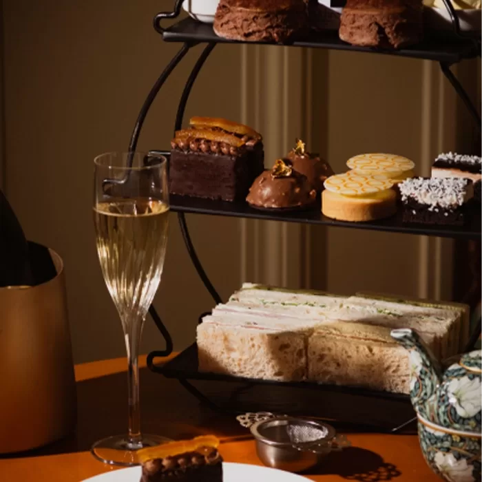 A glass of prosecco on a wooden table next to a black circular cake stand with sandwiches and cakes on 3 tiers.