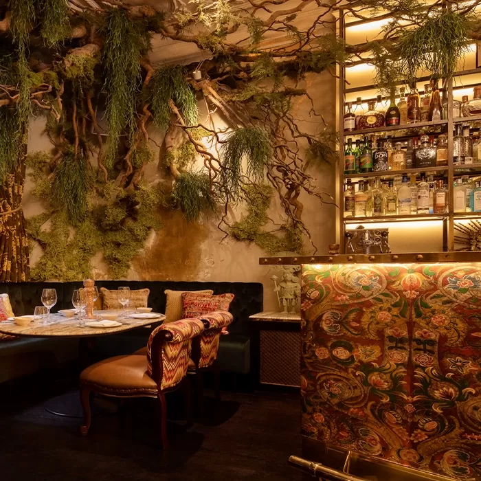 A cosy restaurant interior with lush foliage covering the walls and ceiling. Theres a small table set with glasses and napkins, plush chairs, and a decorative wood-textured bar stocked with various bottles, creating an inviting ambiance. Served at COYA Mayfair.