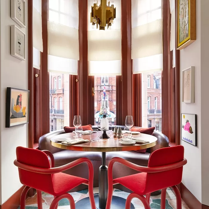 A cosy dining nook with a round wooden table set for two at Mount St Restaurant, Mayfair. It features red chairs and a variety of framed artwork on the walls. Large windows let in natural light, and a modern chandelier hangs above the table.