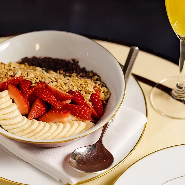 A bowl of sliced bananas, strawberries, granola, and a dark topping sits on a white plate with a silver spoon. A glass of orange juice is beside the bowl, all placed on a cream-colored table of Isabel, Mayfair.