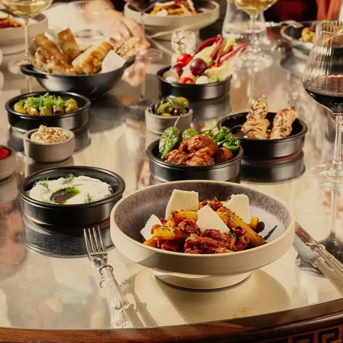 A table filled with a variety of dishes, including a bowl of pasta topped with cheese, small plates of appetizers and dips, and glasses of red and white wine. The table has a reflective surface and the food is elegantly arranged at Bacchanalia, Mayfair.