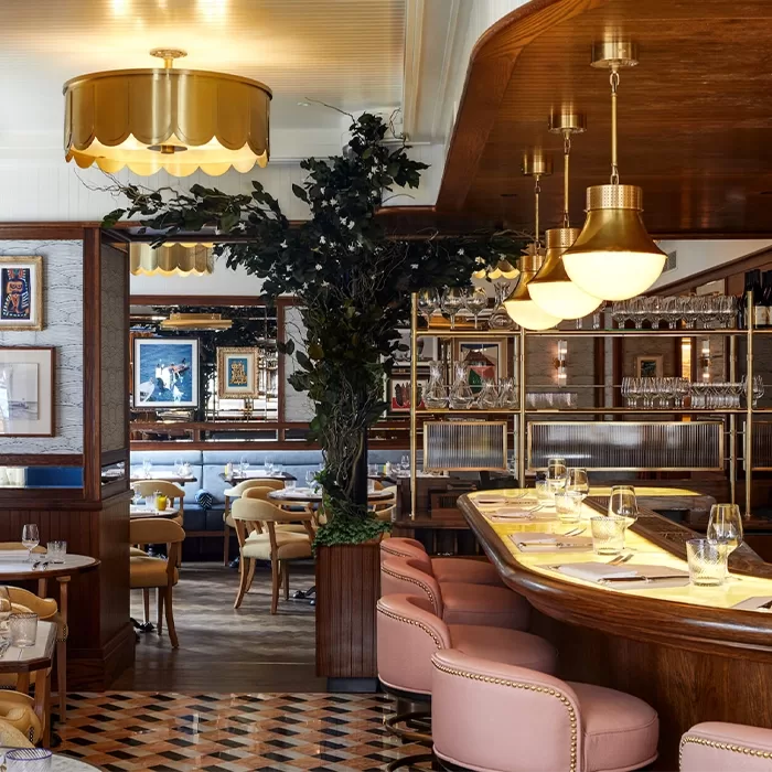 A stylish restaurant interior features a curved bar with pink stools, gold lighting fixtures, and elegantly set tables. The decor includes a large indoor plant, framed artwork on the walls, and a checkered floor pattern at Chucs, Mayfair.