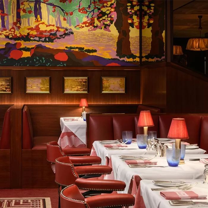A cosy restaurant interior with red leather booths and chairs, white tablecloths, and small red lamps on tables. The walls feature colorful, impressionistic art depicting a vibrant landscape. The setting is warm and inviting at The Colony Grill Room at The Beaumont, Mayfair.