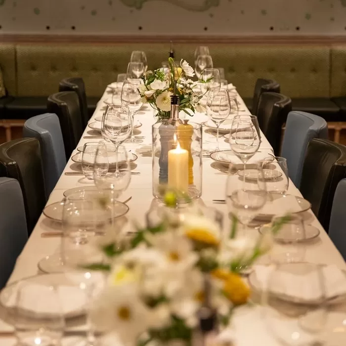 A long dining table is elegantly set with white plates, wine glasses, and decorative vases of white and yellow flowers. A lit candle in a glass holder is centered, with plush green and blue chairs lining either side of the table at Socca Mayfair: Petit Salon.