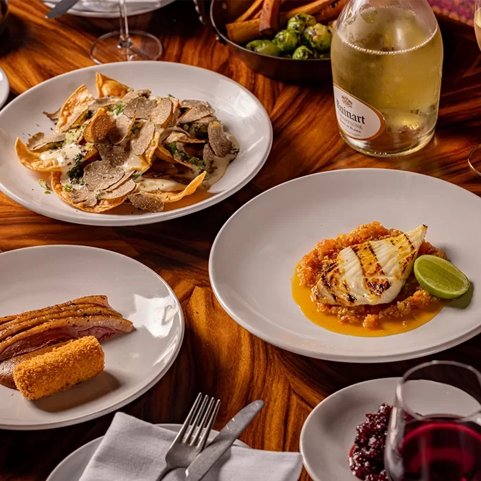 A table with various dishes including truffle-topped pasta, grilled fish with sauce and lime, a meat dish with a croquette, and a bottle of Ruinart champagne. Additional plates and a glass of wine are also visible, served at Decimo at The Standard, Kings Cross in London.