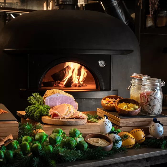 A cosy setting featuring a wood-fired oven with an open flame. In the foreground, theres a spread of cooked ham, bread, jars of condiments, greens, and colorful bowls on a table adorned with fresh herbs and brussels sprouts, served at Ekstedt at the Yard, Great Scotland Yard in London.