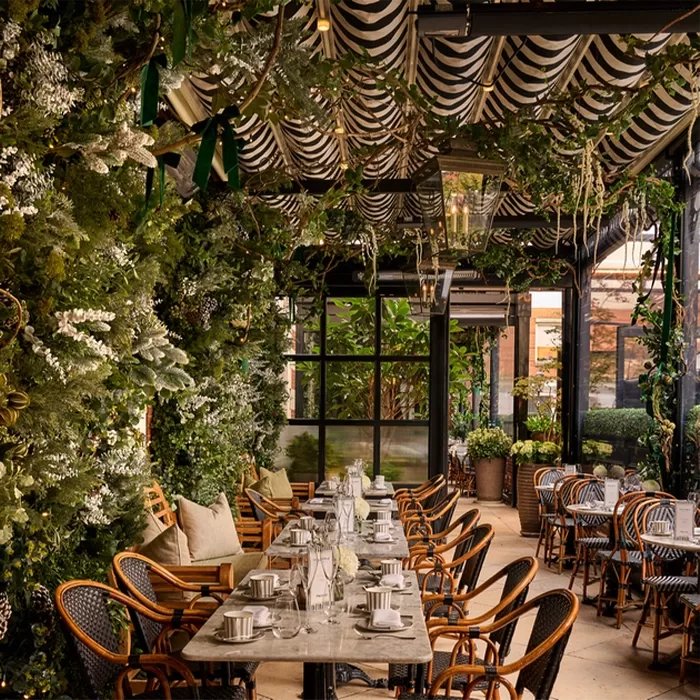 A cosy outdoor dining area features a long table set for a meal, surrounded by wicker chairs. Lush greenery and plants hang from the ceiling and walls, creating a garden-like atmosphere under striped awnings. Warm lighting enhances the ambiance of Dalloway Terrace, Bloomsbury in London.