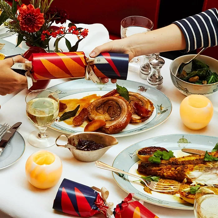 A festive dinner table with two people pulling a Christmas cracker. Plates feature roasted meat, vegetables, and a side dish. The table is decorated with candles, flowers, and a glass of white wine at Ham Yard Hotel, Soho in London.