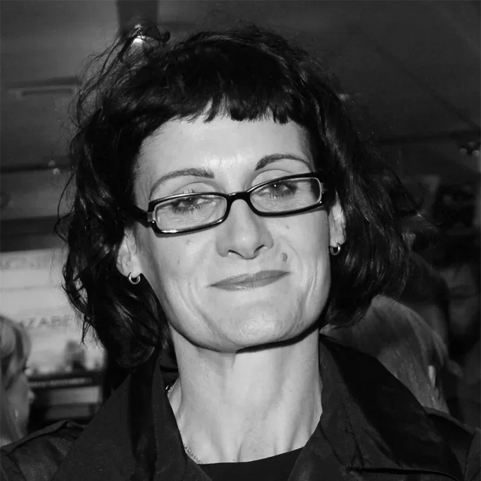 A black and white photo of Alexandra Byrne wearing glasses and a jacket. The background is blurred, focusing on their facial expression.
