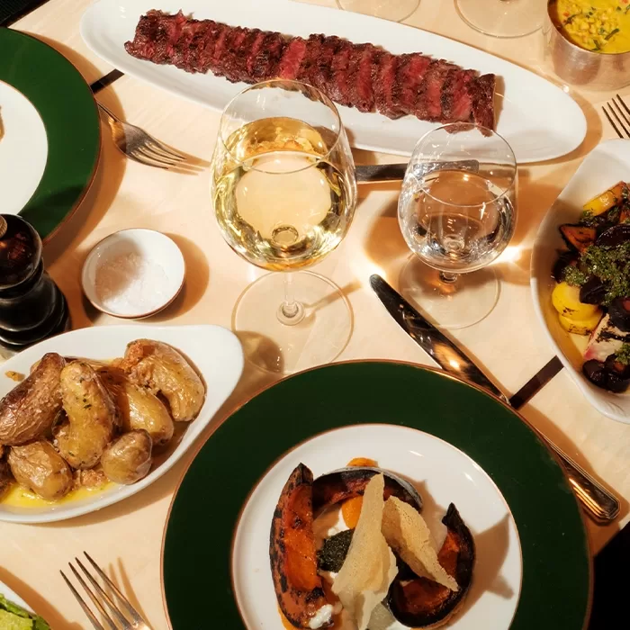 A table set with an array of dishes: sliced steak on a platter, roasted potatoes, squash with crisp toppings, a dish of roasted vegetables, and glasses of white wine and water. The table is elegantly arranged with cutlery and a pepper mill.
