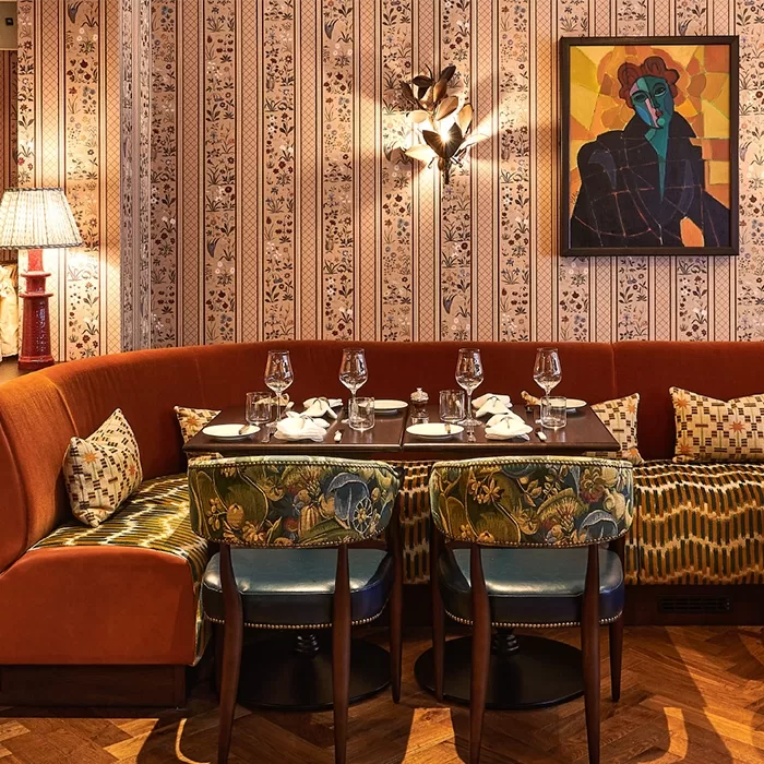 Vintage restaurant interior with a rich, patterned wallpaper, an orange velvet curved sofa, floral cushions, and a dark wooden table set for four. A colorful abstract painting and a unique wall lamp enhance the cozy, stylish ambiance.