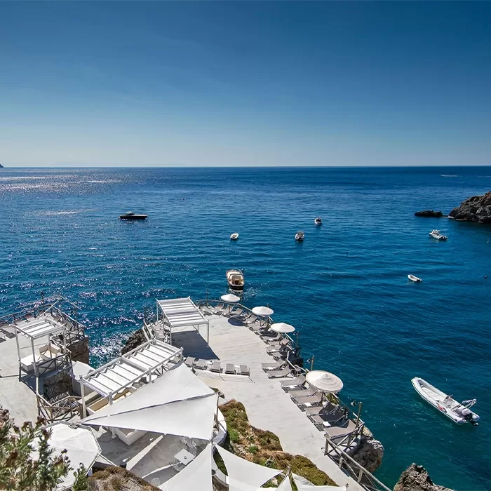 A seaside resort with white sun loungers, umbrellas, and shaded areas on rocky terraces overlooking a calm, blue sea with several small boats floating nearby under a clear sky.