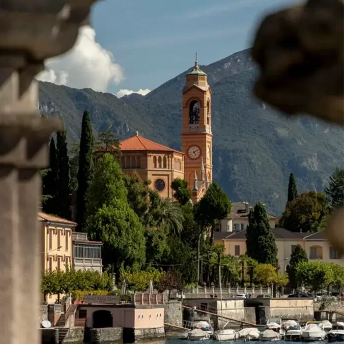 A scenic view of a lakeside village with a tall clock tower, surrounded by lush greenery and mountains in the background. Boats are docked along the waterfront under a clear sky.