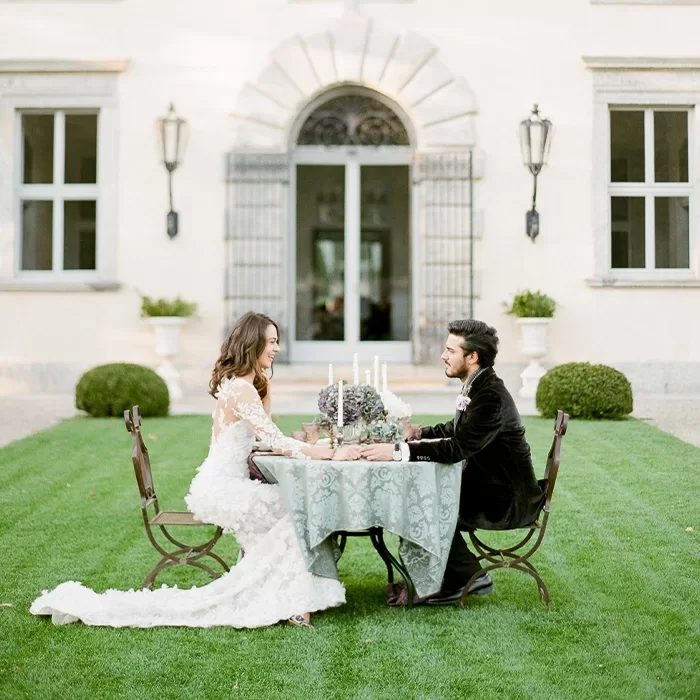 A bride and groom sit across from each other at a small table set with candles and flowers on a lawn in front of an elegant building, holding hands and smiling.