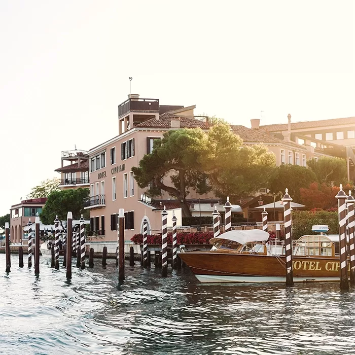 A wooden motorboat is docked by striped poles on a canal beside Hotel Cipriani, with trees and pastel buildings in the background under a bright sky.