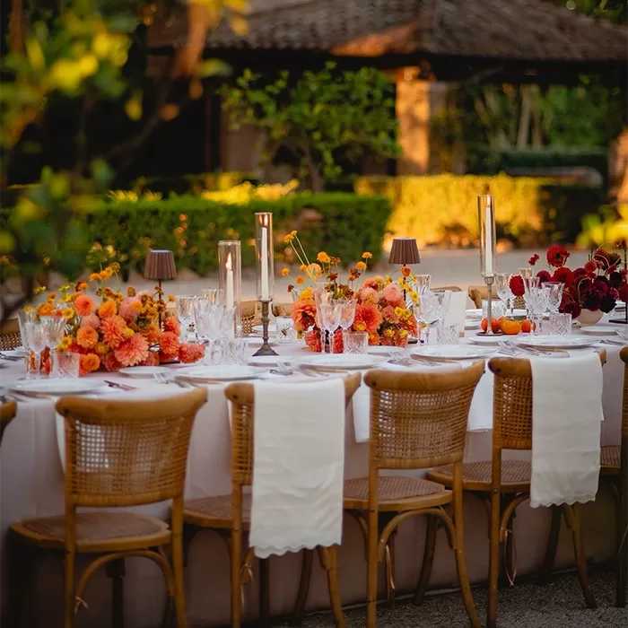 A long outdoor dining table with wicker chairs, white linens, elegant glassware, and floral centerpieces in orange and red hues is set for an event in a lush garden at sunset.