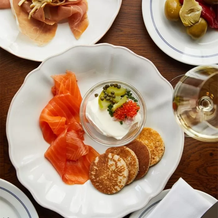 A plate with smoked salmon slices, small pancakes, and a glass dish of cream topped with capers and fish roe. Surrounding plates hold cured meats and marinated vegetables. A glass of white wine is also visible.