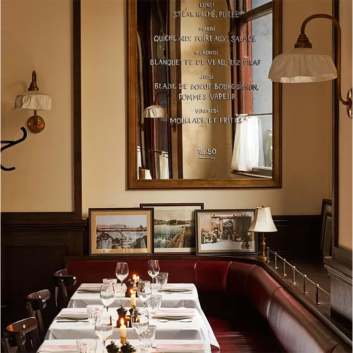 Elegant restaurant interior with neatly set tables, lit candles, cushioned red bench seating, framed photos against the wall, a large mirror displaying a French menu, and warm wall-mounted lamps.