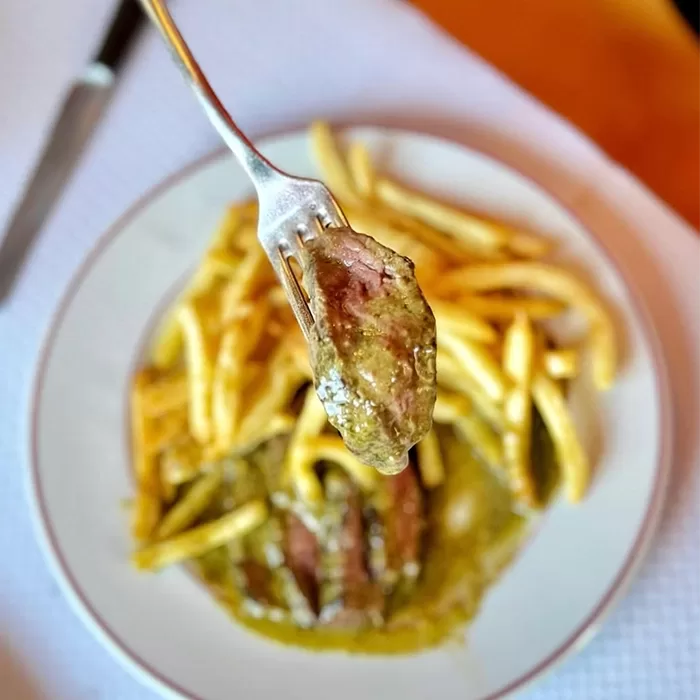 A close-up of a fork holding a piece of steak with green sauce, above a plate of steak, green sauce, and golden French fries.