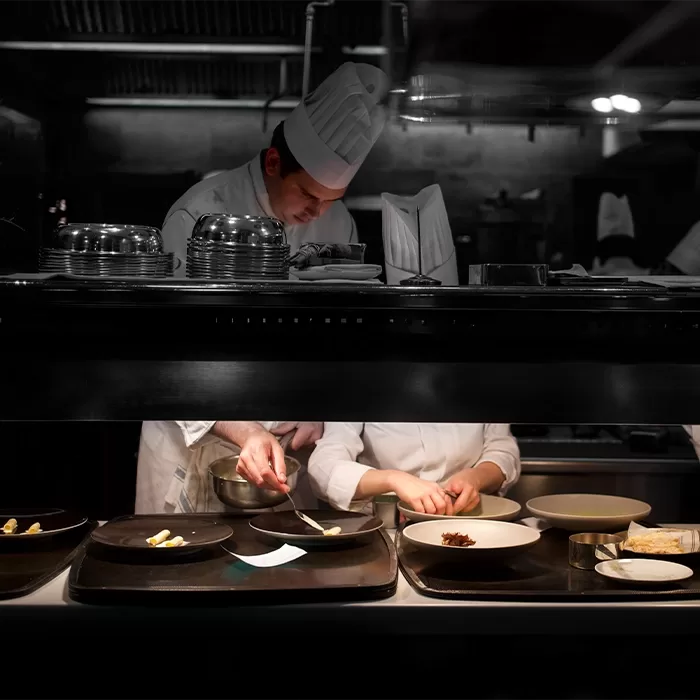 A chef in a white uniform and tall hat prepares food in a dimly lit restaurant kitchen, plating dishes alongside another chef whose face is mostly obscured. Various plates of food are lined up on the counter.