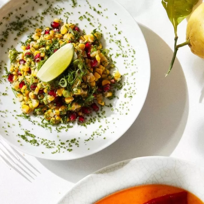 A white plate with a colorful corn salad garnished with herbs, pomegranate seeds, and a lime wedge, set on a white table next to a yellow fruit and part of another plate in the corner.