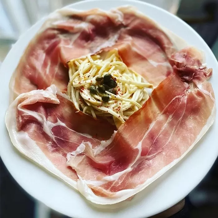 Thin slices of prosciutto arranged in a circle on a white plate, with a small nest of spaghetti topped with capers and seasoning in the center.
