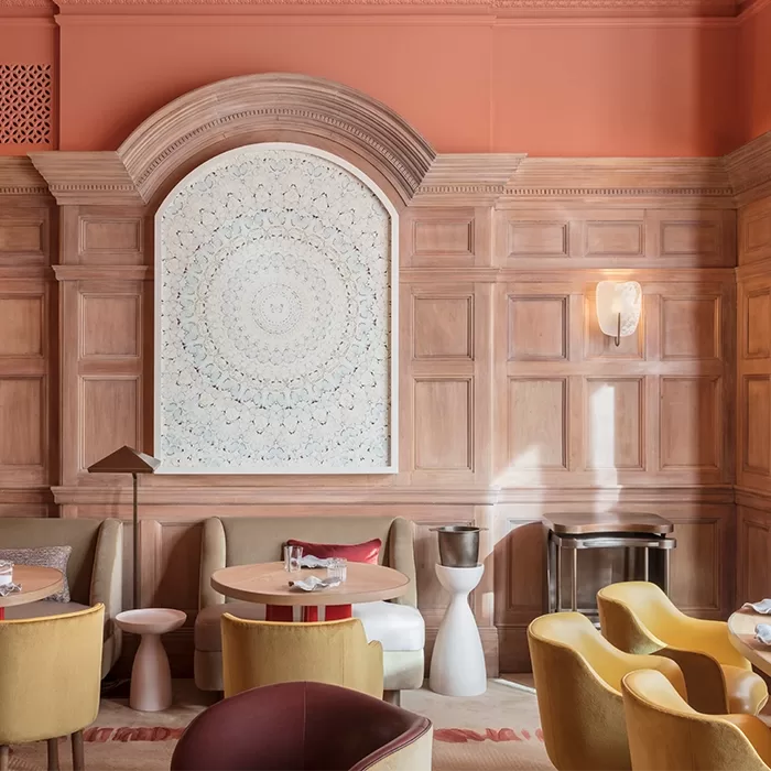 Elegant dining room with wood paneling, coral-colored walls, and a large, intricate white artwork framed by an arched molding. Modern furniture includes round tables, beige sofas, and yellow chairs.