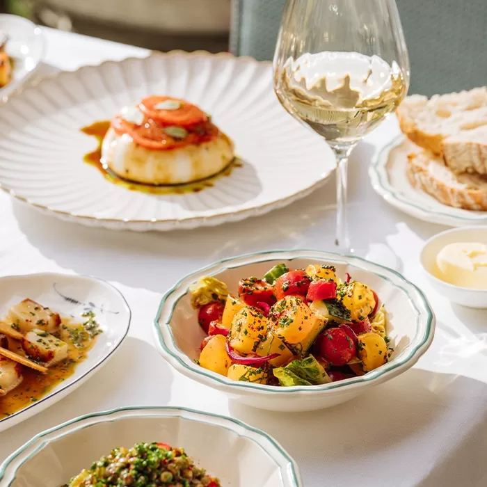 A table set with dishes including a bowl of colorful tomato salad, a plate with a molded appetizer topped with sauce, sliced bread, a glass of white wine, and other small plates of food on a white tablecloth.