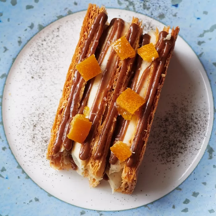 A close-up of a dessert with flaky pastry layers, cream filling, chocolate drizzles, and small orange candied fruit pieces, served on a white plate with a dusting of black powder on a blue surface.
