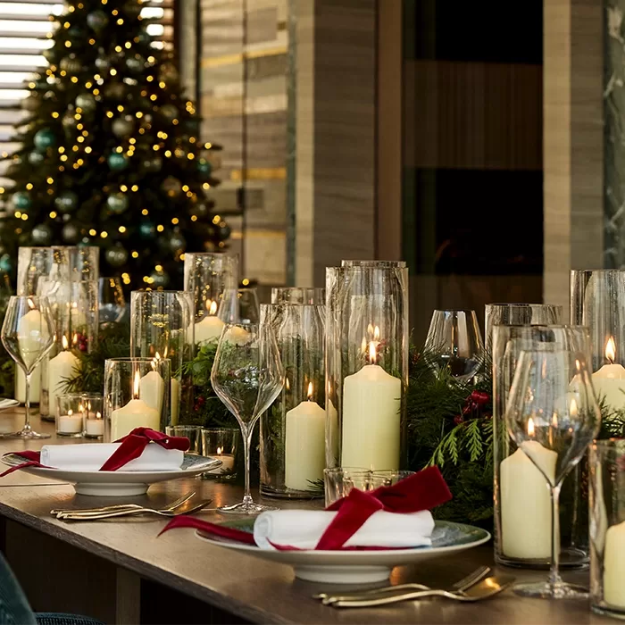 A festive dining table is set with tall glass candle holders, white candles, plates, napkins with red ribbons, and greenery. A decorated Christmas tree with lights is visible in the background.