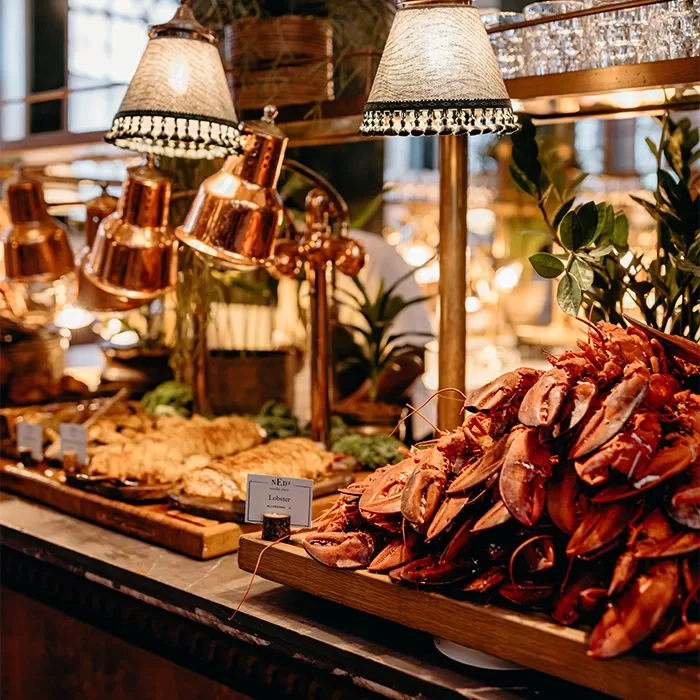 A buffet table displays a large pile of lobster claws and other seafood under warm lighting, with decorative plants and heat lamps in the background. A card labeled Lobster is visible among the seafood.